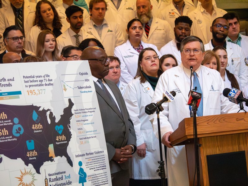 An older man in a doctor's coat speaks at a podium, many more people in doctor's coats behind him. A sign to the left reads 2024 Birth and Death Trends with markings all over a US Map