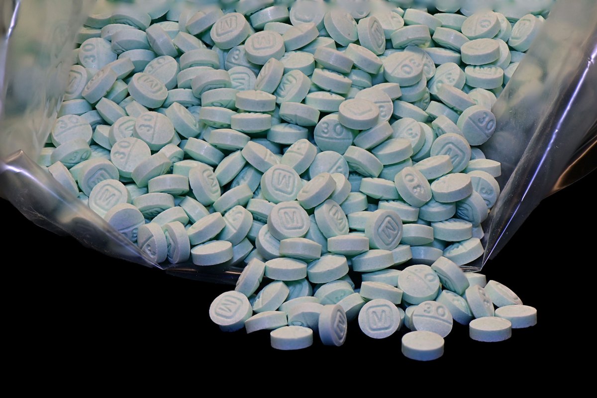 A bag of light blue pills with the impression of an M on one side spill out of a clear bag