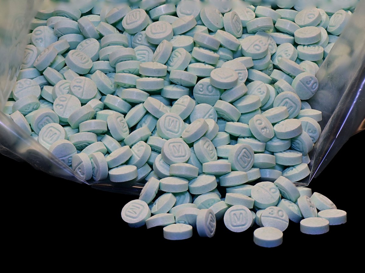 A bag of light blue pills with the impression of an M on one side spill out of a clear bag