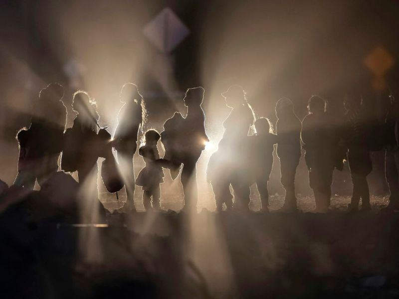 Profiles of people in a line, lit from behind by car lights