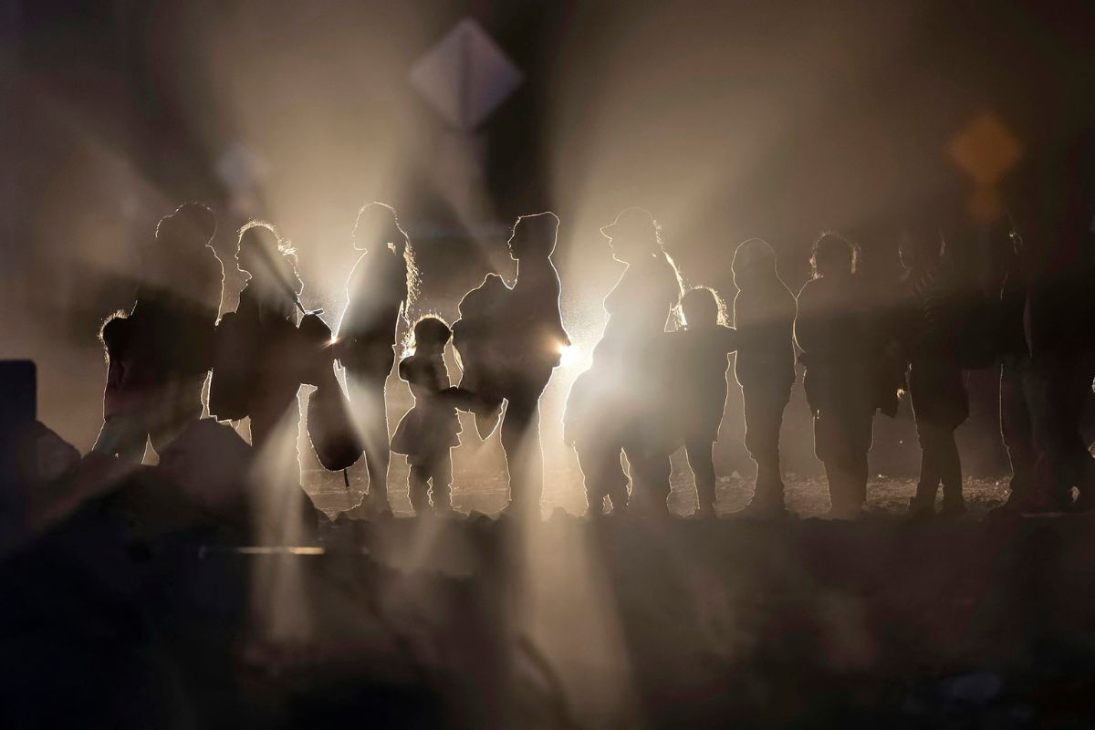 Profiles of people in a line, lit from behind by car lights