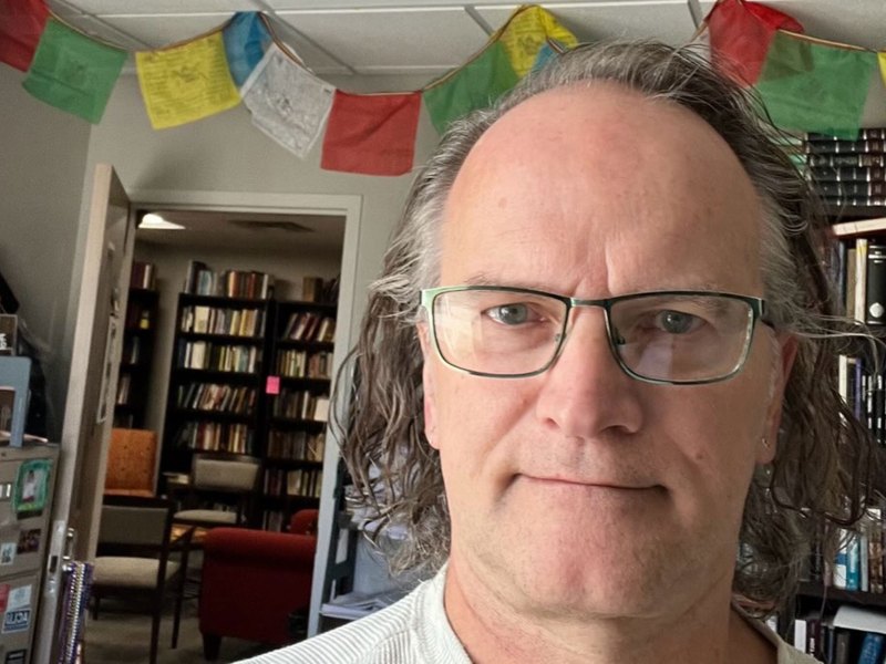 A man in glasses with slightly longer hair stands in an office with colorful prayer flags hanging from the ceiling, and very busy bookshelves in the background