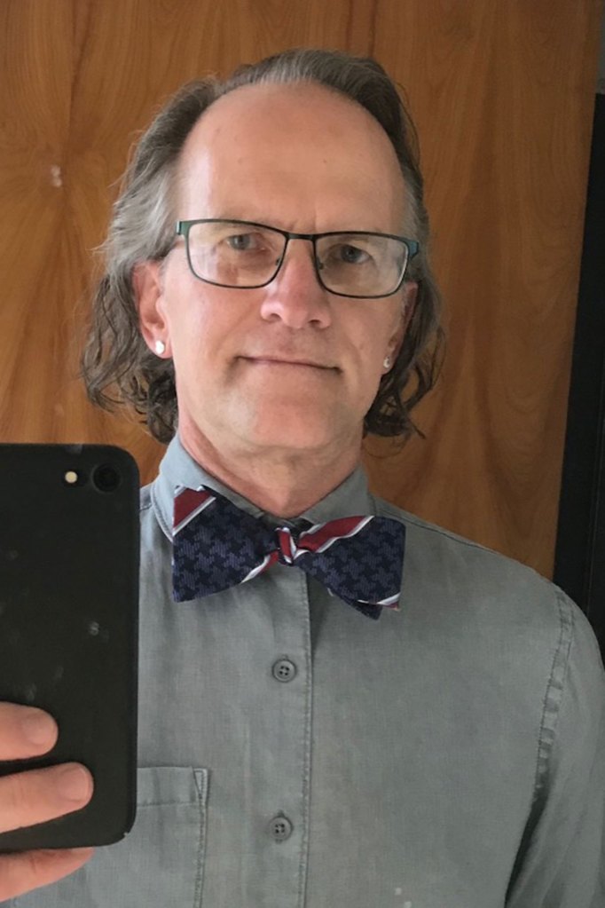 A man in glasses with slightly longer hair, wearing a grey button up shirt with a blue and red striped bowtie, takes a selfie