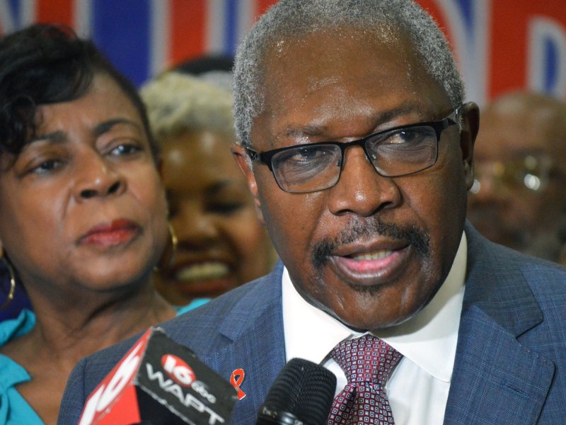 A man in glasses and a suit speaks int a reporter's mic as his wife and crowd look on behind him