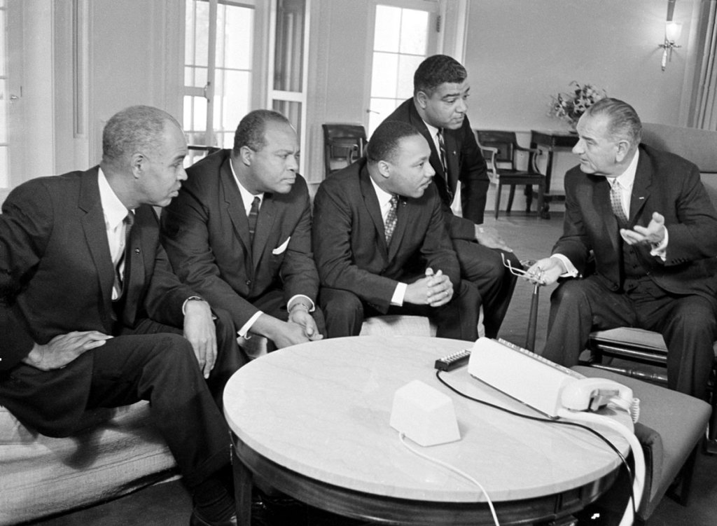 An old black and white photo of a white man on the right side, speaking to four black men seated opposite of him