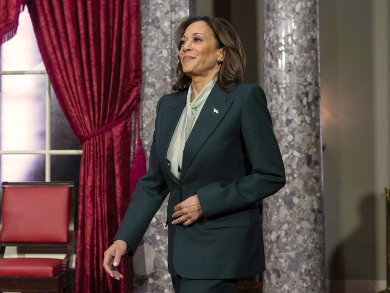 A woman in a suit walks into a room with marble columns and red velvet curtains
