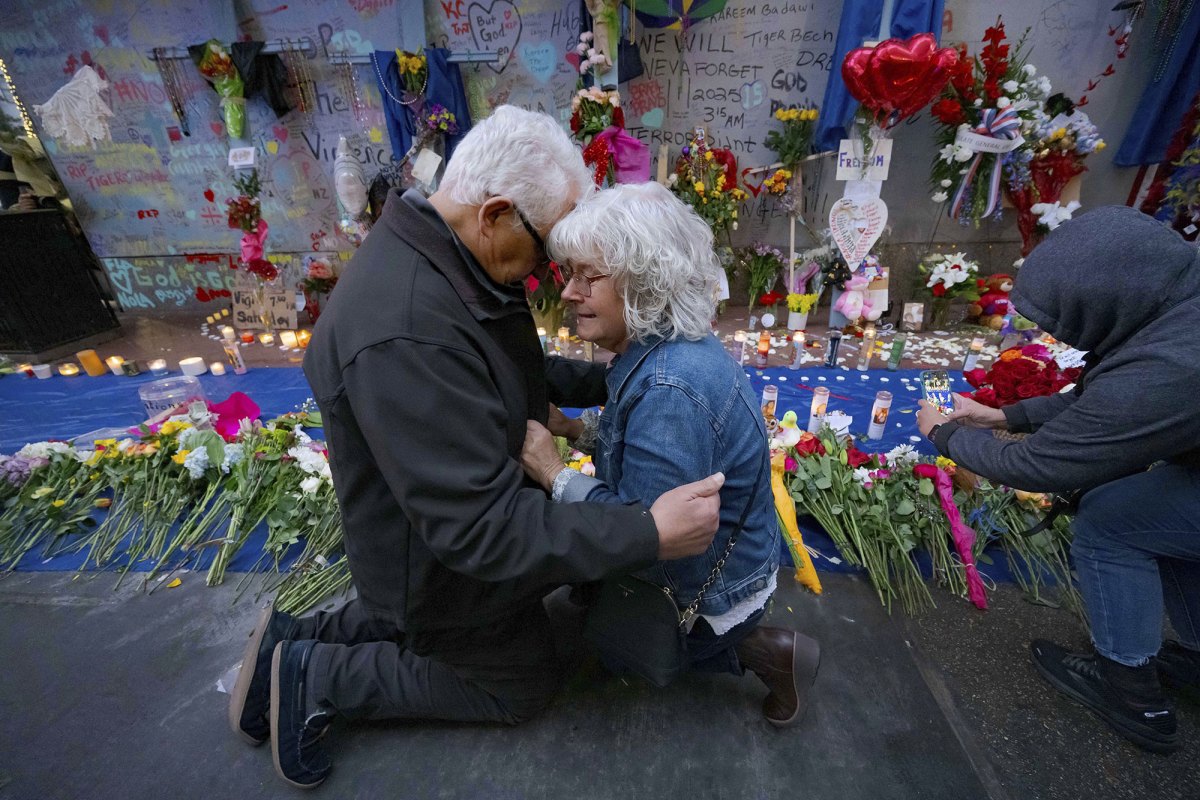 Two older people kneel and embrace in front of a street memorial covered in flowers and crosses