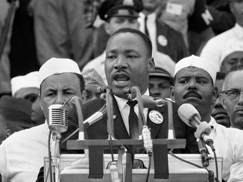 An old black and white photo of a black man speaking at a podium with lines of men behind him