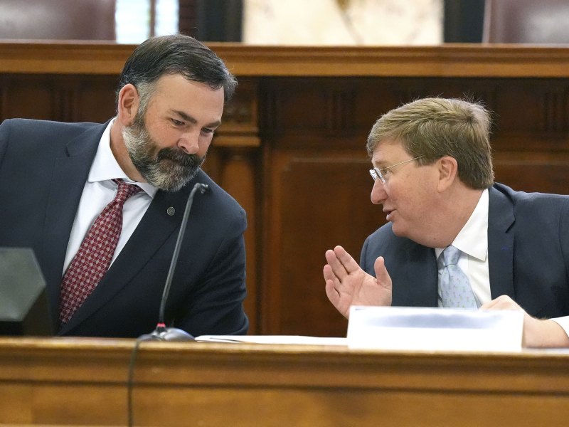 Two men in suits lean in behind a desk and talk