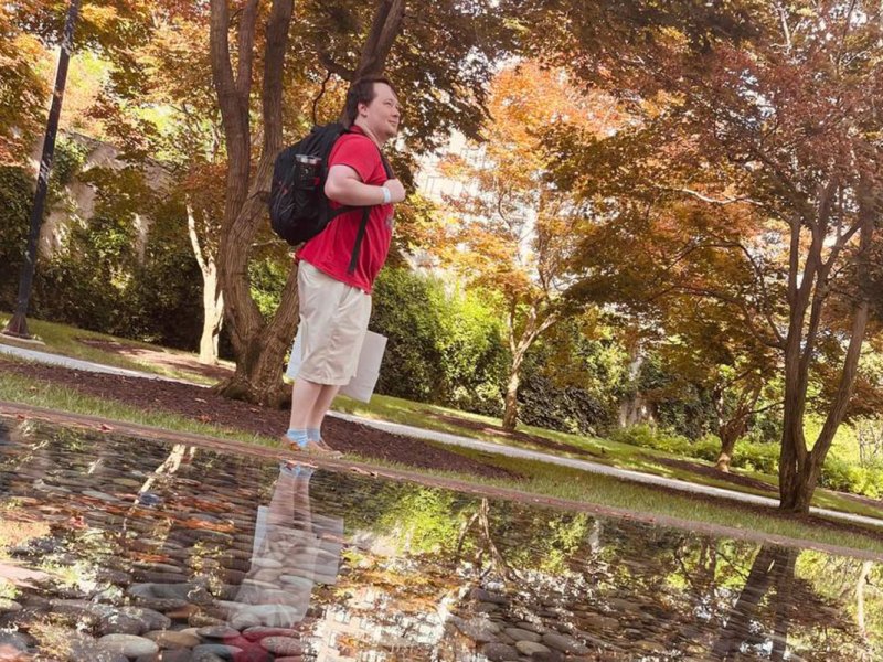 A man in a red top and wearing a backpack poses beside a reflection pool