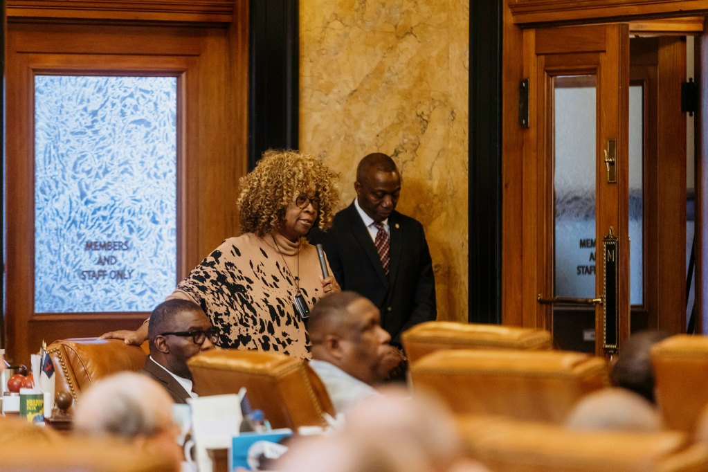 A man and woman stand near the back of a room, listening