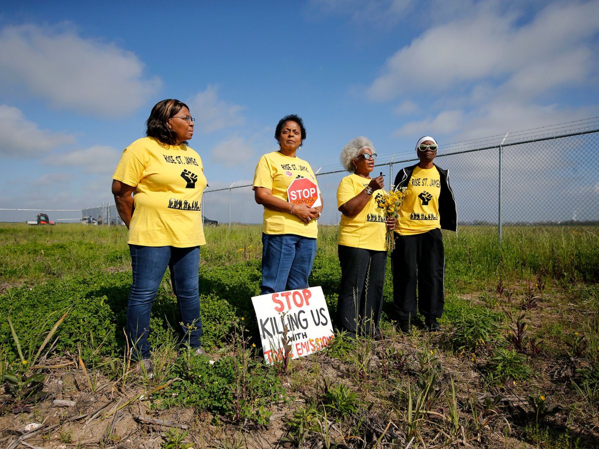 Four women in matching yellow tshirts that say RISE St. JAMES stand outside in a field beside a fence with a sign that says STOP KILLING US WITH EMISSIONS