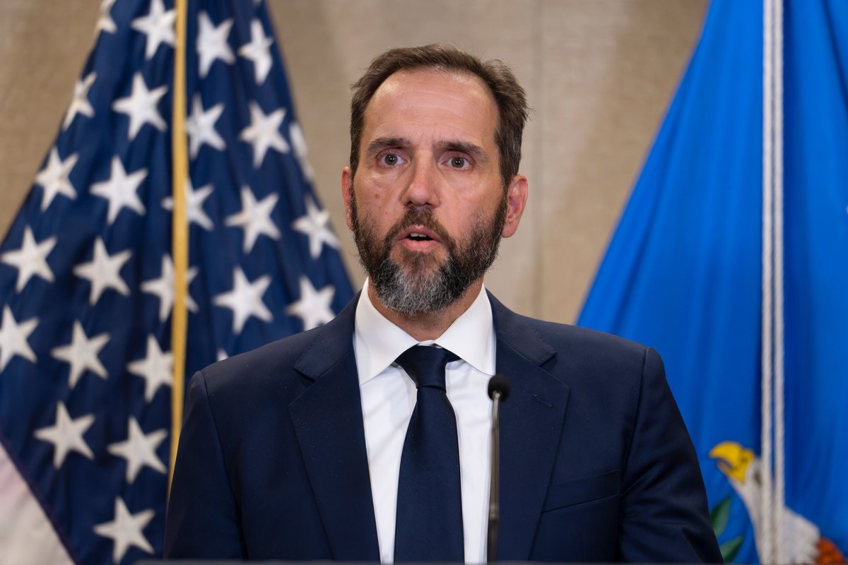 A bearded man in a suit speaks in front of a US flag