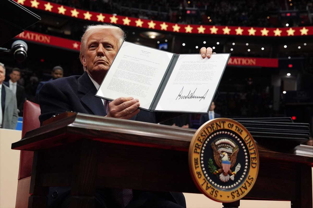 A man sitting on a large stage holds an opened two page paper folio with a large signature