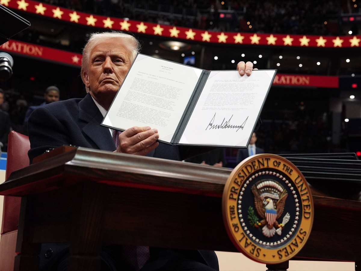 A man sitting on a large stage holds an opened two page paper folio with a large signature