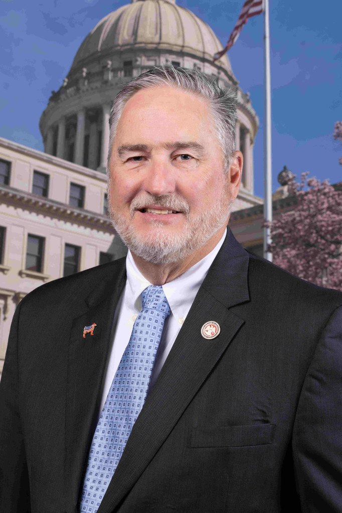 Official portrait of a man in a black suit posed in front of the capitol building