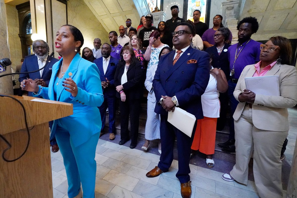 A woman in a light blue suit speaks at a podium. Many people stand behind her and listen on.