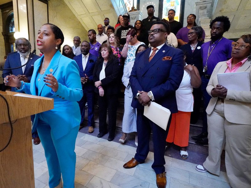 A woman in a light blue suit speaks at a podium. Many people stand behind her and listen on.