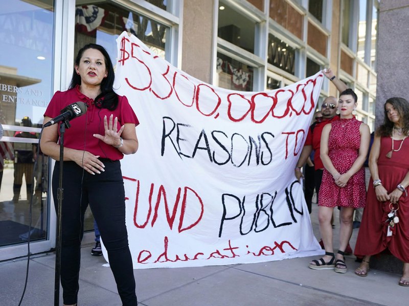 People in red outfits present outside beside a large sign that reads $5,300,000,000 reasons to FUND public education