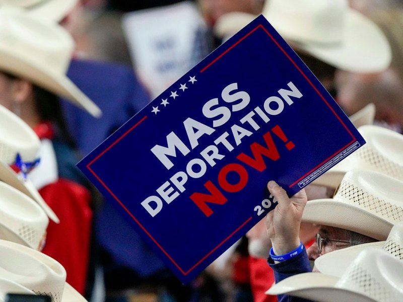 A hand in a crowd of people wearing matching white cowboy hats, the sign reads Mass deportation NOW!