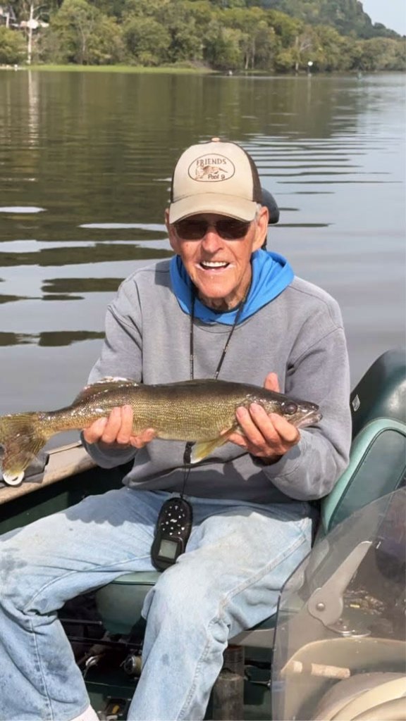 An older man sits on a boat on the water holding a fish in his hands