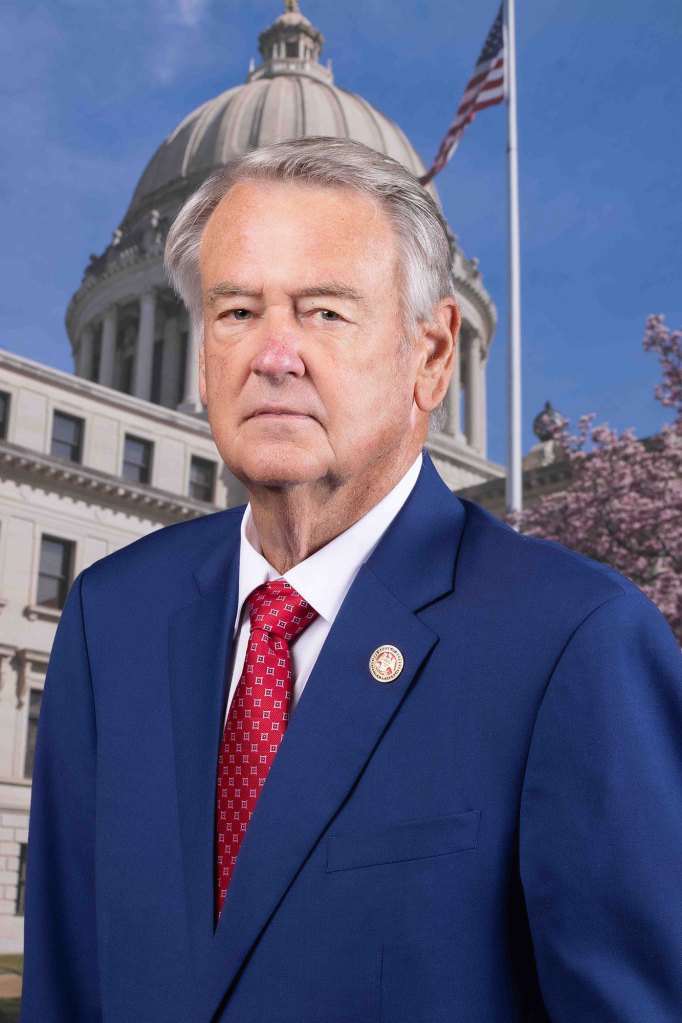 Official portrait of a man in a blue suit with red tie, posed in front of the capitol building