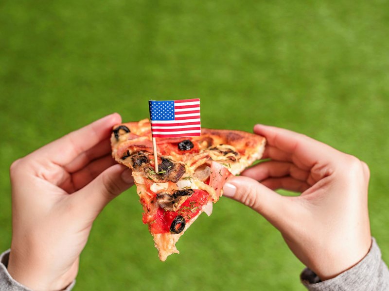 A pair of hands holds up a slice of pizza against a green background. A little US Flag is staked in the center of the slice.