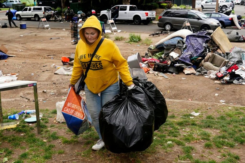 A woman in a yellow hoodie carries her belongings in trash bags with destroyed tents visible in the background