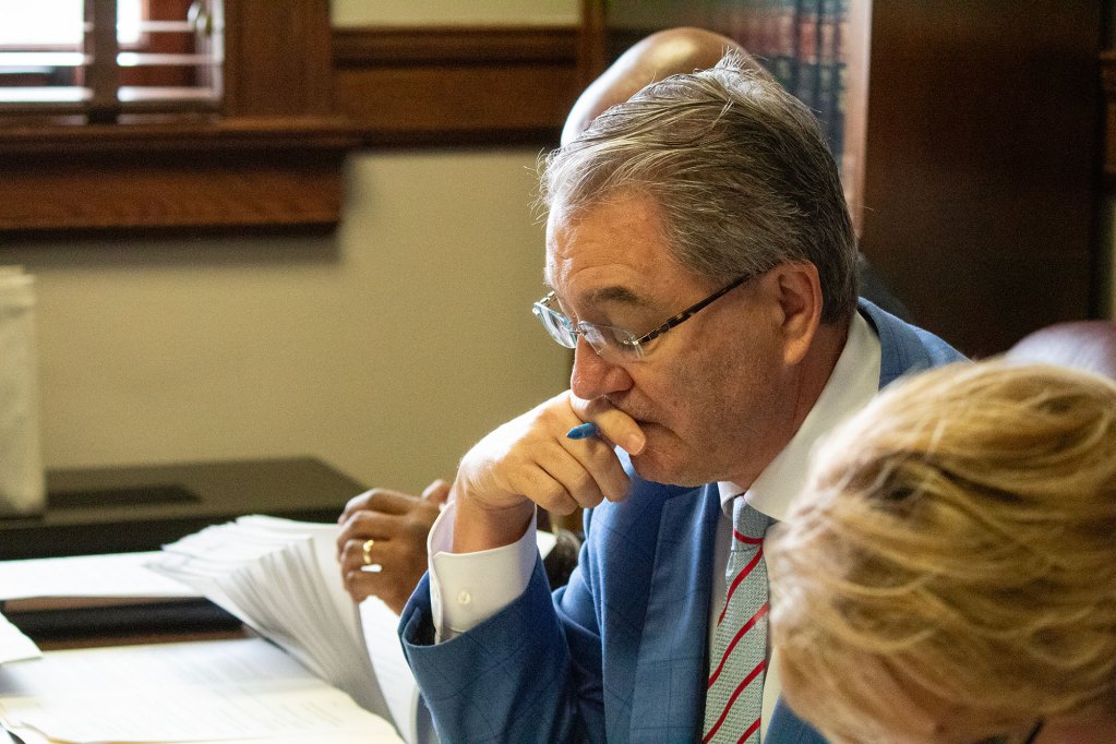 A man in glasses holds a blue pin in a hand that he rests his face on as he looks at paperwork on the desk before him