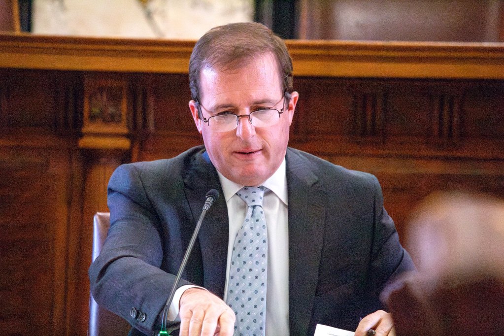 A man in a grey suit and light blue dotted tie sits and speaks in front of a small mic