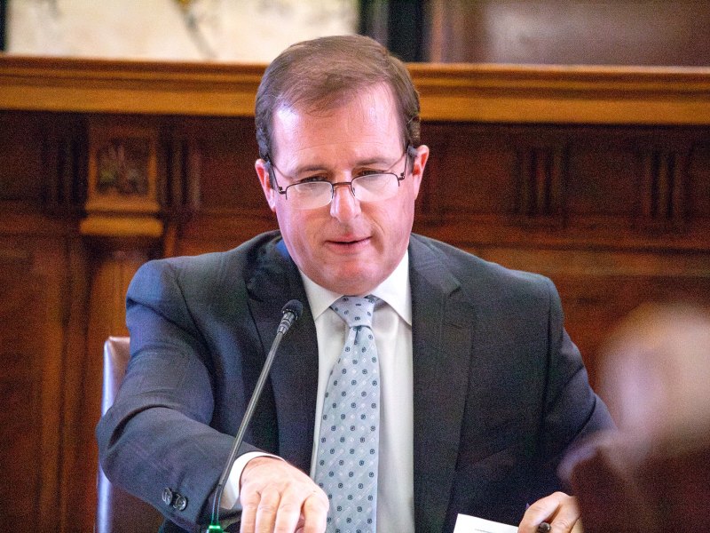 A man in a grey suit and light blue dotted tie sits and speaks in front of a small mic