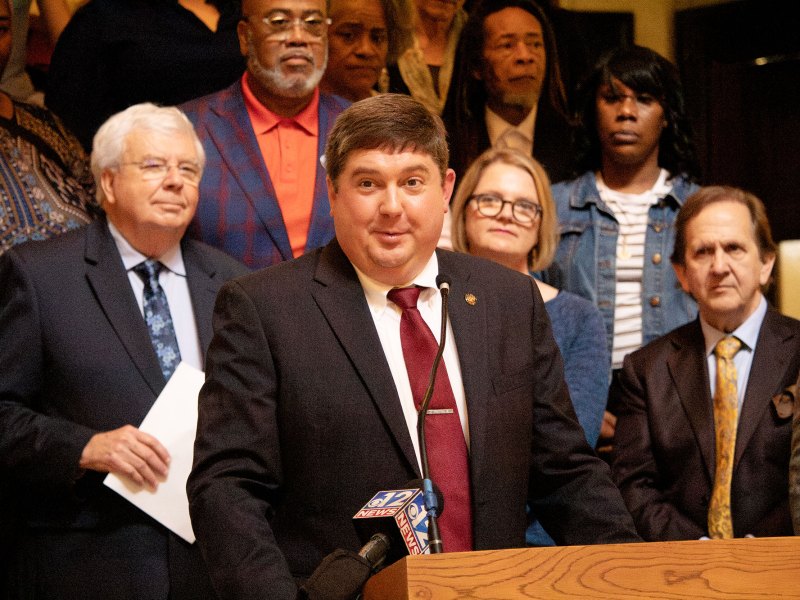 A man in a suit speaks at a podium as others look on behind him