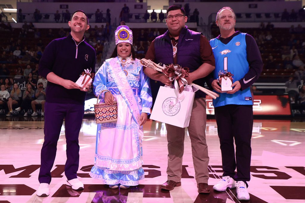 Four people line up on a basketball court with gifts in their hands. The woman is dressed in native blues and white.