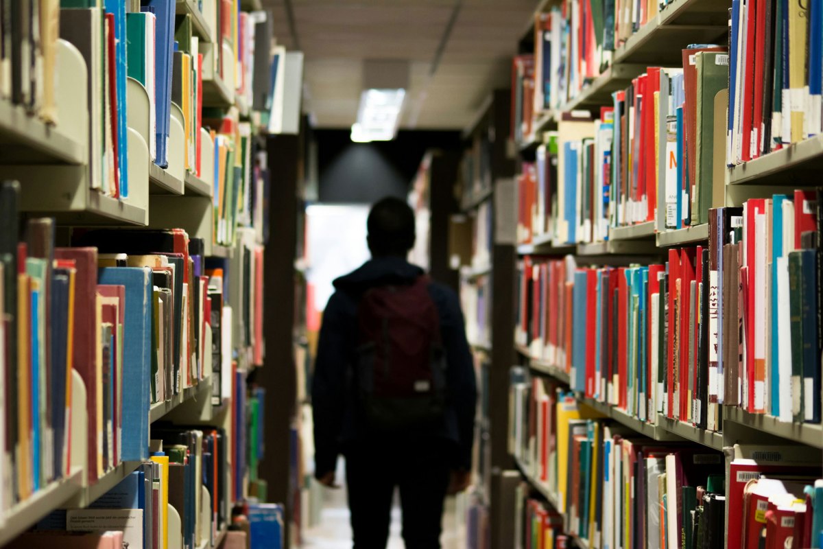 A shadowed student with backpack on walks between rows of bookshelves