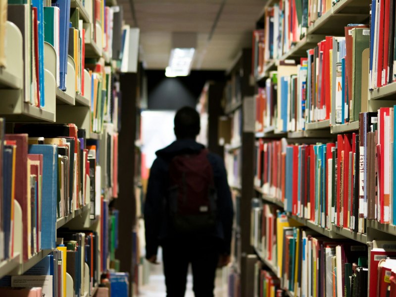 A shadowed student with backpack on walks between rows of bookshelves