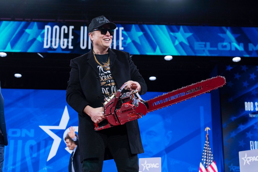 A man in sunglasses and all black clothes wields a red chainsaw on a blue political stage