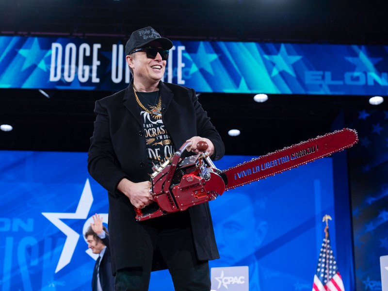 A man in sunglasses and all black clothes wields a red chainsaw on a blue political stage