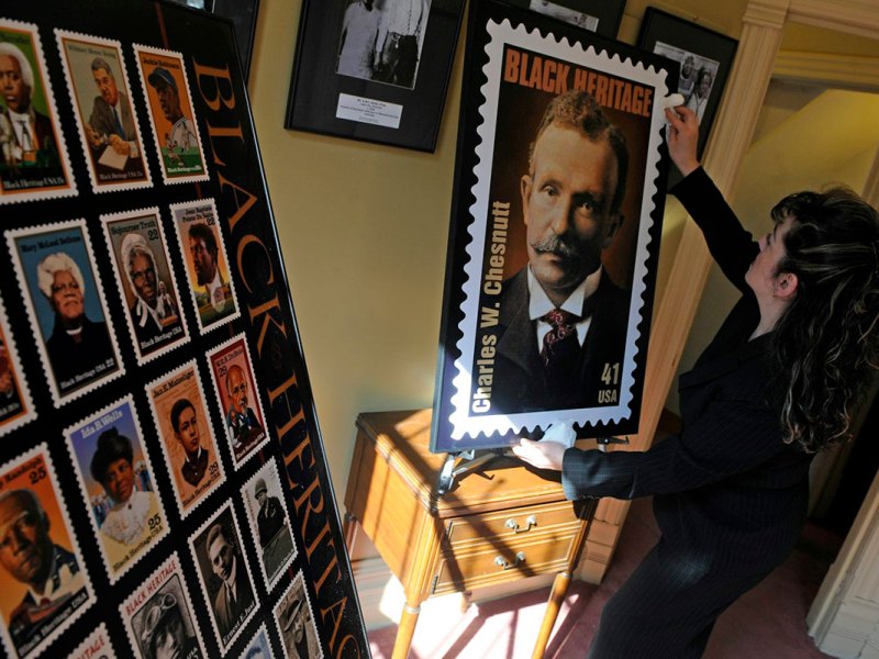 A woman holds up a a blown up photo of a stamp that reads Black Heritage, Charles W. Chestnutt