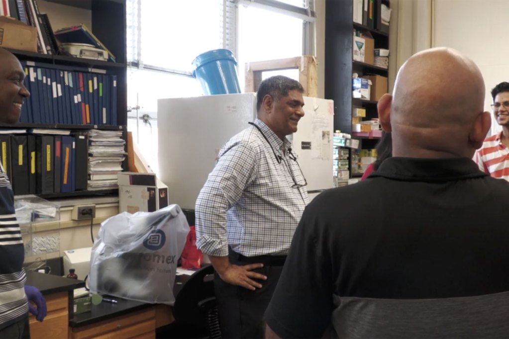 Four men are talking together in a lab. The back wall is covered in shelves