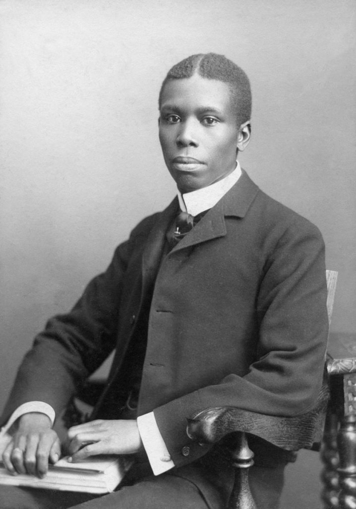 A young black man dressed a dark suit sits in a chair with a book and pen resting in his lap