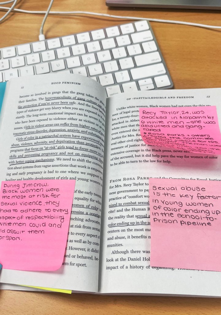 An open book sitting on top of a keyboard, sentences seen underlined and notes added on pink sticky notes.
