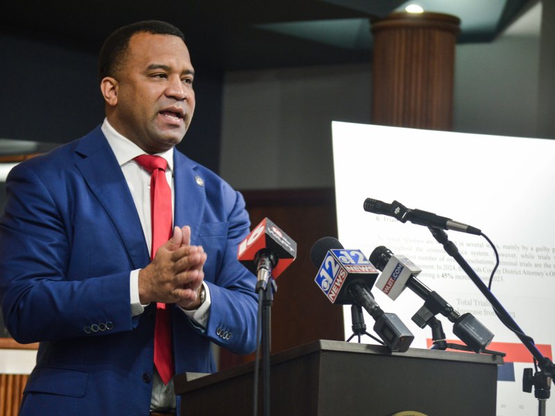 A man in blue suit and red tie speaks with hands held together behind a podium covered in press microphones