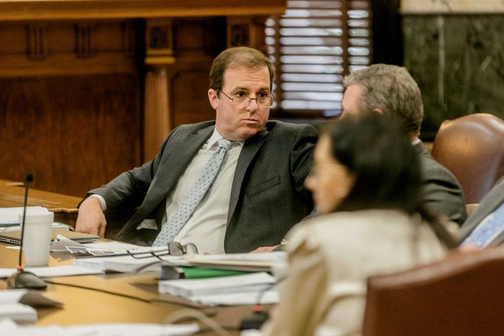 A man in glasses leans back at a legislative meeting towards other people tables at desks covered in paperwork
