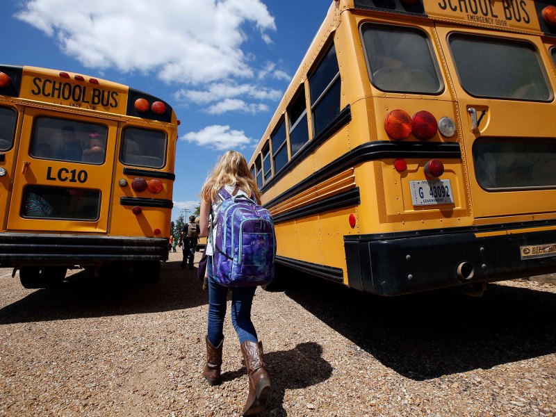 A child wearing a blue backpack walking between two orange school busses