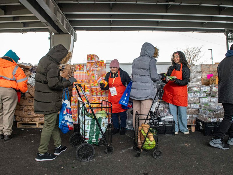 Three people in coats walk through a pop up food pantry that appears to be set up under a bridge. Workers in orange aprons hand packages of groceries out.