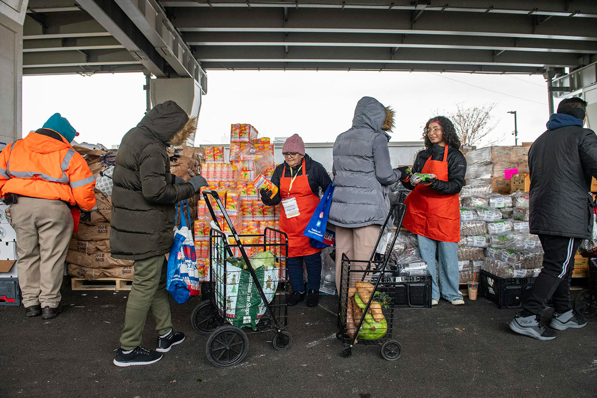 Three people in coats walk through a pop up food pantry that appears to be set up under a bridge. Workers in orange aprons hand packages of groceries out.