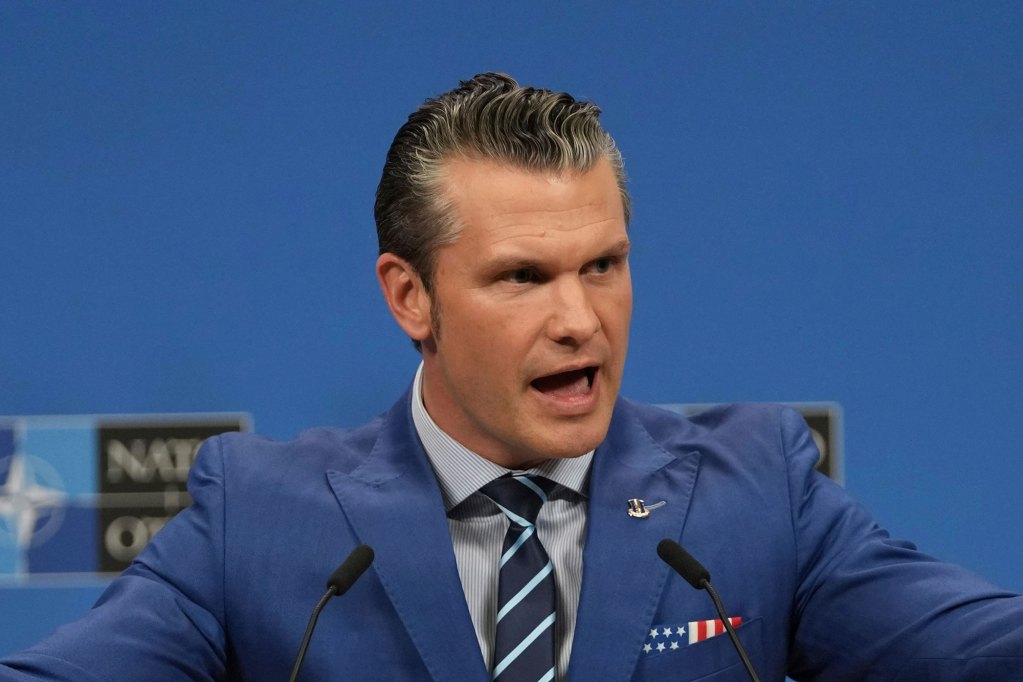 A man in a blue suit with US Flag pocket square, speaking in front of a NATO backdrop