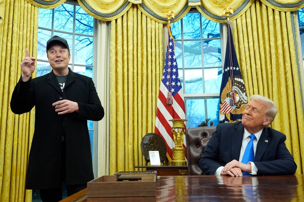 Two men are inside in a room with golden curtains. One stands and talks while pointing upwards as the other, seated at a desk, smiles smugly