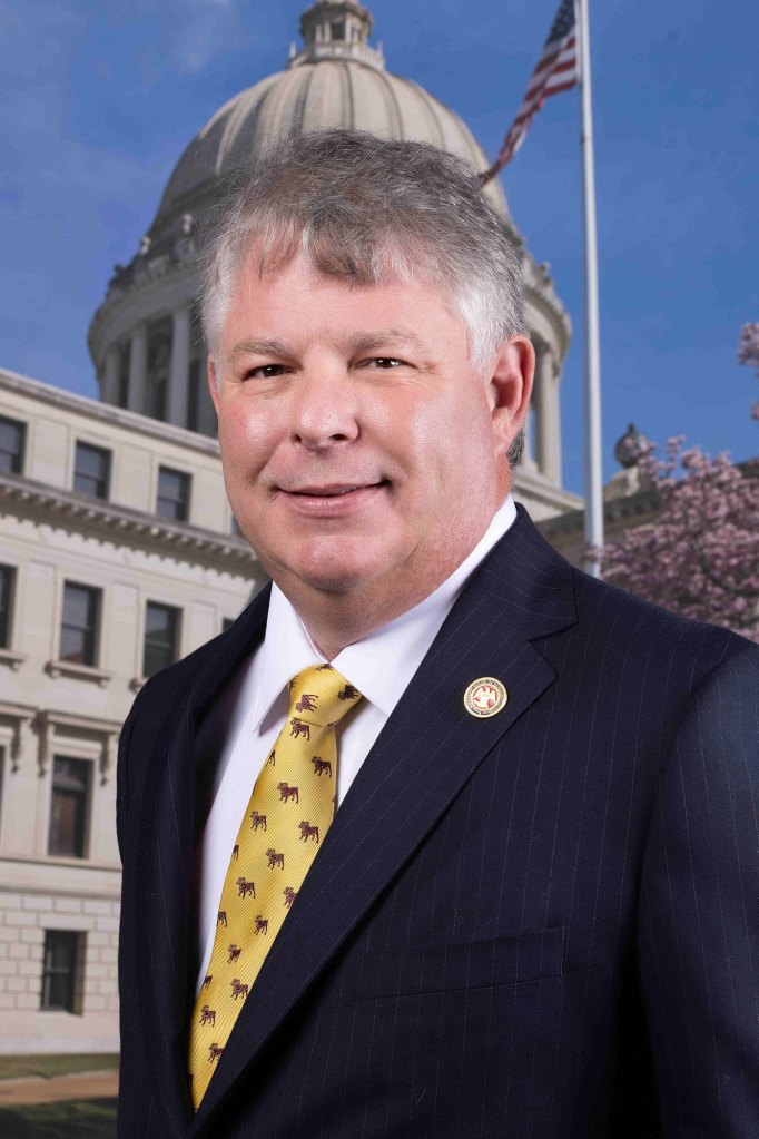 An official portrait of a man in a black suit and gold tie with small repeating pattern