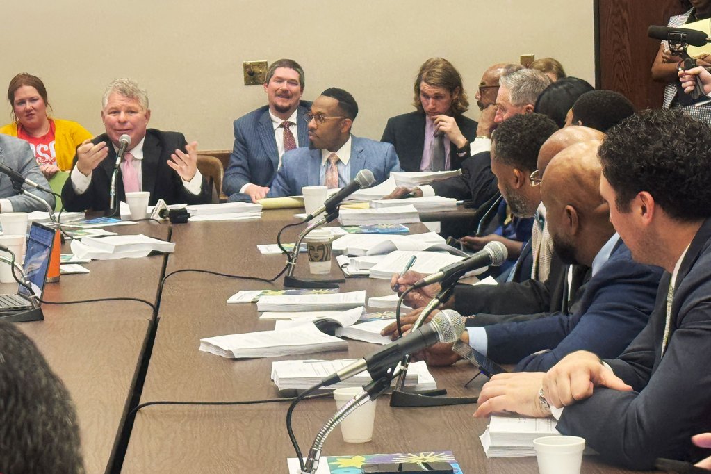 A group of people seated around a large, brown table. Placed on the table are microphones and several stacks of paper.